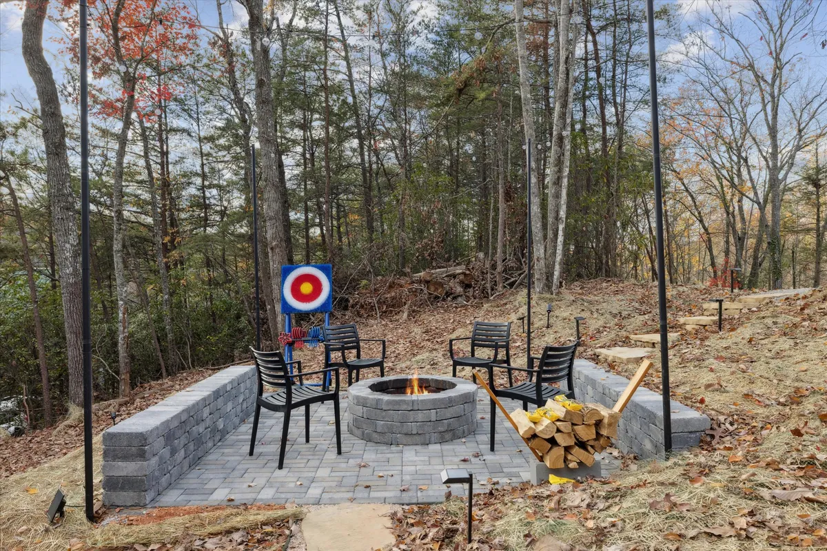 This image features a cozy and adventurous courtyard with a prominent fire pit, surrounding seating area, an archery (axe throwing) target, and a neatly stacked wood pile. The space is framed by trees and rustic elements, capturing a sense of camaraderie and outdoor fun.