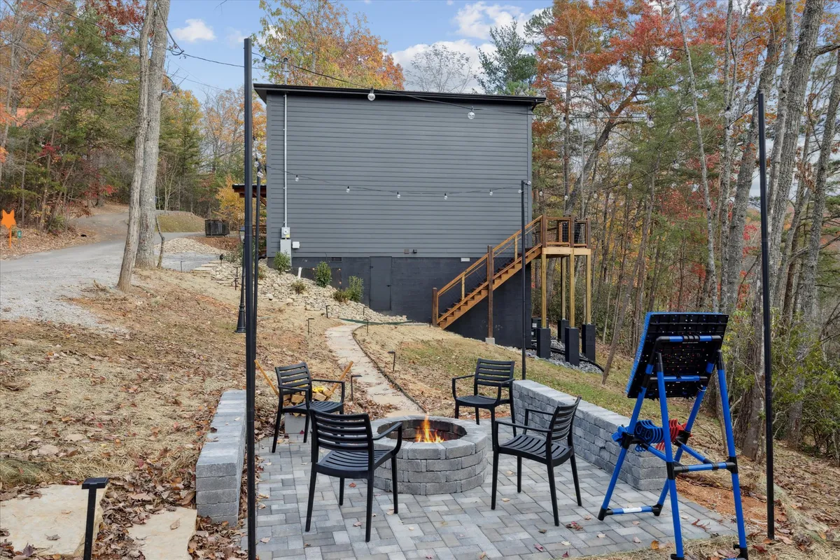 An inviting fire pit encircled by comfortable seating sits in a natural setting with a pathway and a wooded backdrop. The image conveys a relaxed environment ideal for storytelling, making s’mores, or stargazing in the evening light.