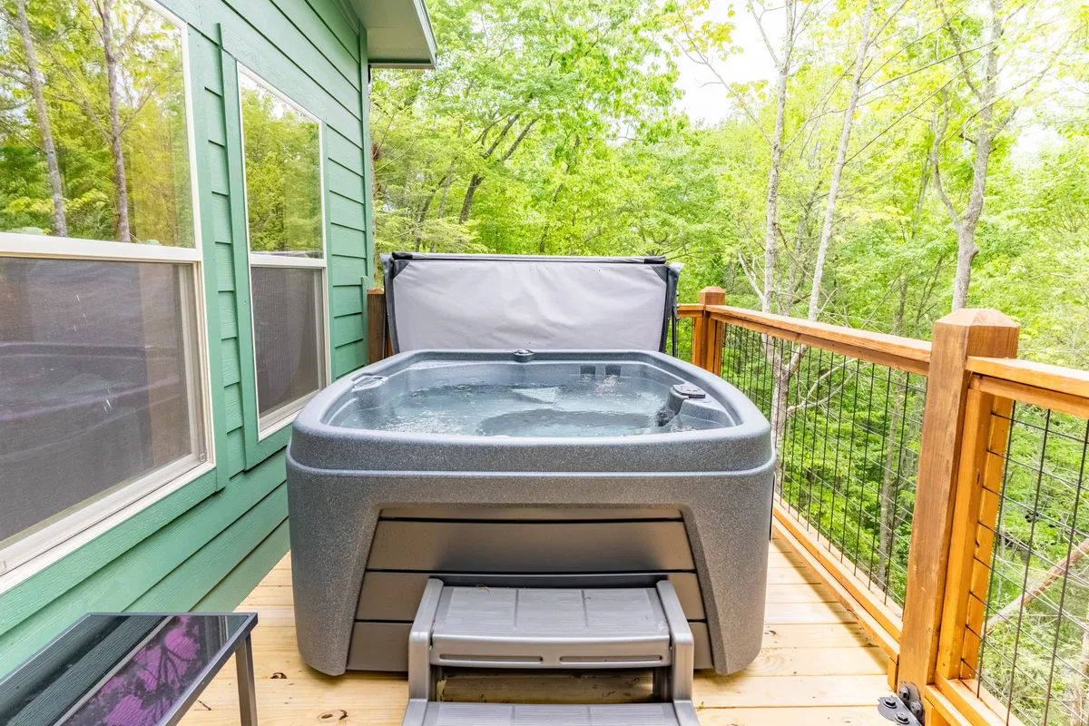 A wooden deck with a hot tub overlooks dense, leafy trees, suggesting a peaceful retreat after a day outdoors. The inviting hot tub is surrounded by forest, and the deck offers space for social evening gatherings or quiet relaxation.