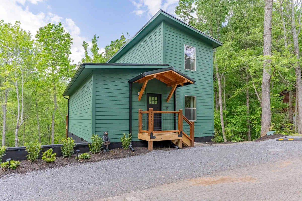 The image features the green exterior of two inviting cabins nestled among lush trees, each with a charming wooden porch perfect for relaxing. The natural surroundings evoke a sense of peace, with the porches offering views ideal for morning coffee or evening gatherings under the stars.