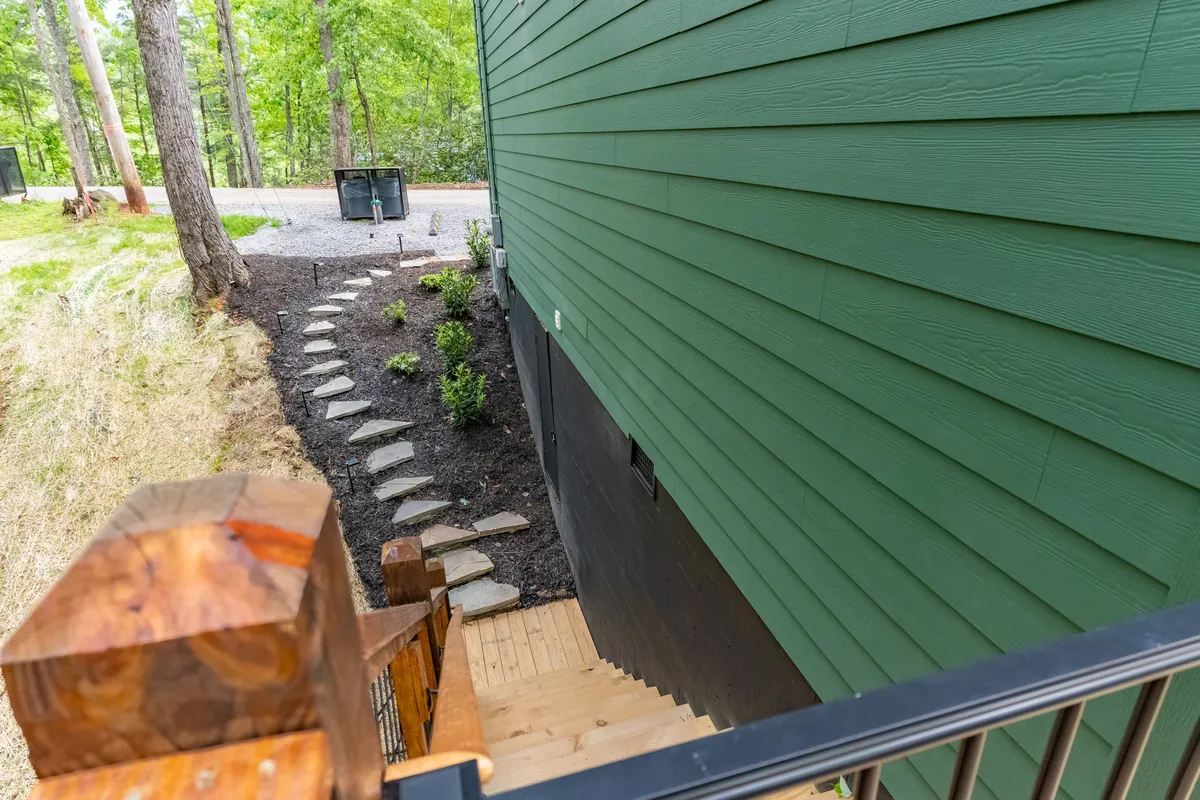 A tranquil stone pathway leads to a green-sided cabin, with wooden stairs and abundant greenery. The outdoor layout hints at a fire pit and space for group activities, reflecting the safe, communal feel ideal for reunions and celebrations.