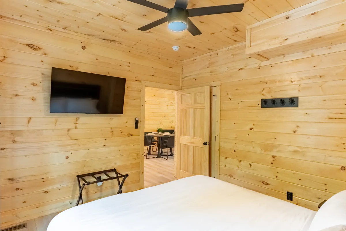 A cozy bedroom featuring a bed with crisp white bedding, a wall-mounted TV, and warm wooden walls. The space is bathed in soft light, ideal for relaxing, watching a movie, or unwinding with a book.