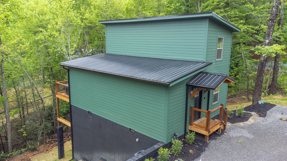 The green exterior of one of the cabins is shown with a welcoming wooden porch set amid lush natural surroundings. The porch invites guests to relax, with the forest as a peaceful backdrop—ideal for stepping right outside to enjoy fresh air and friendly conversation.